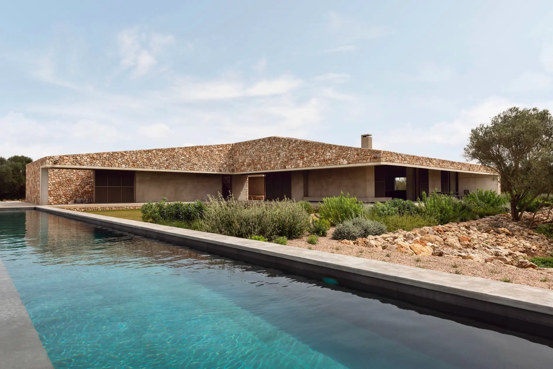 A modern stone-clad finca with minimalist architecture, surrounded by Mediterranean landscaping, featuring an infinity-style swimming pool in front.