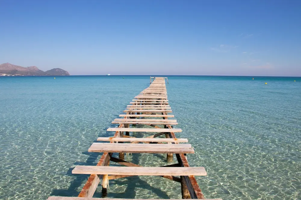 Wooden pier at Playa de Muro in Mallorca, an iconic and most-photographed spot on the island, stretching into the turquoise Mediterranean Sea.